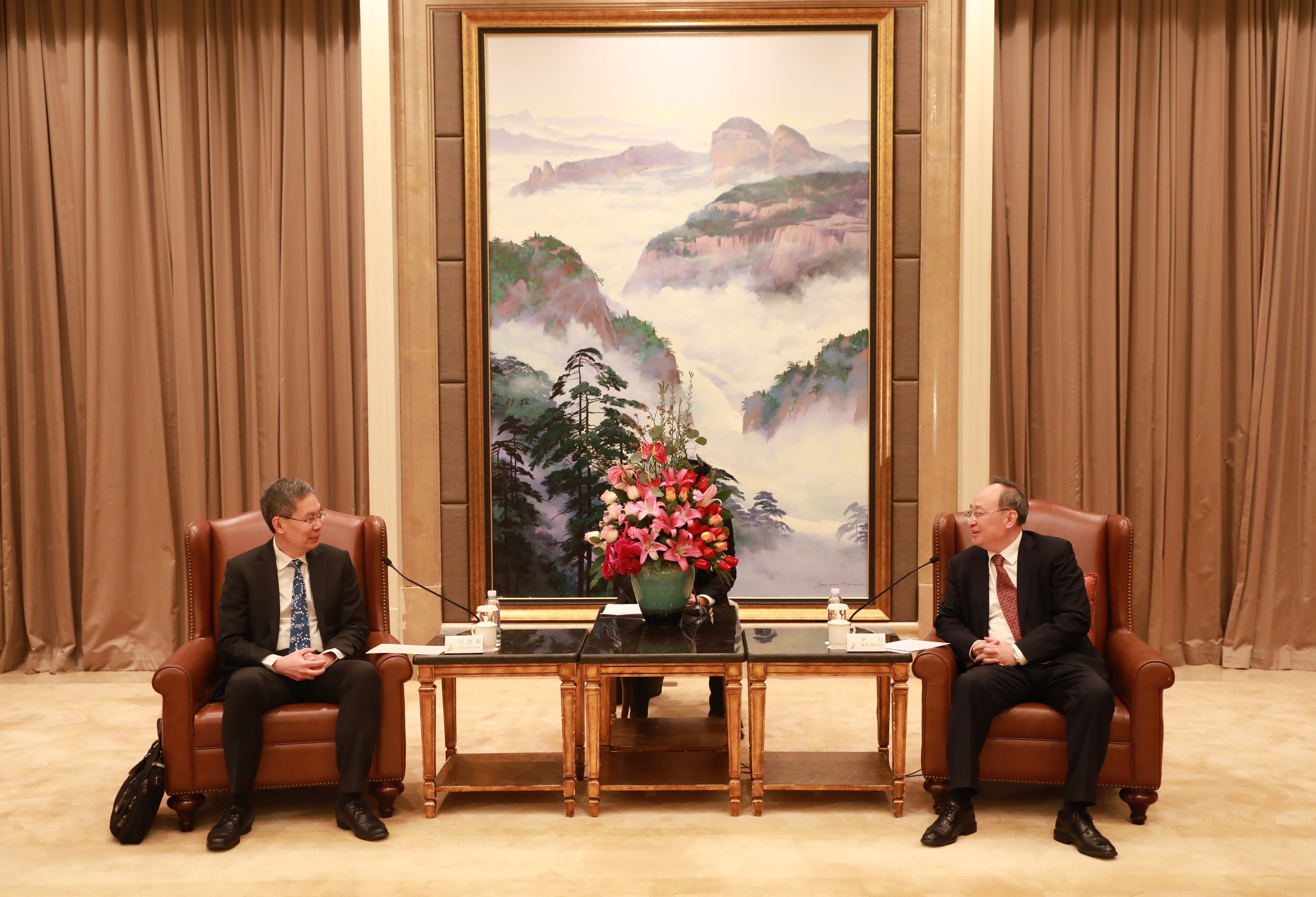 Two men in suits sit facing each other in leather chairs before a landscape painting.